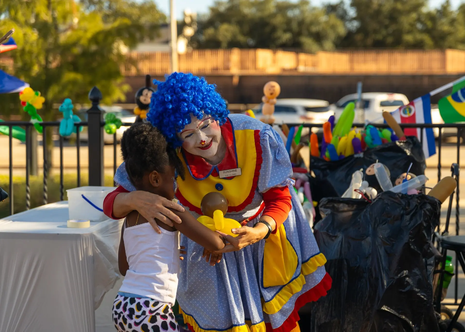 A clown in a blue wig hugs a child beside a balloon stand, photographed by AVO Dynamics at Mobile Latin Fest 2023