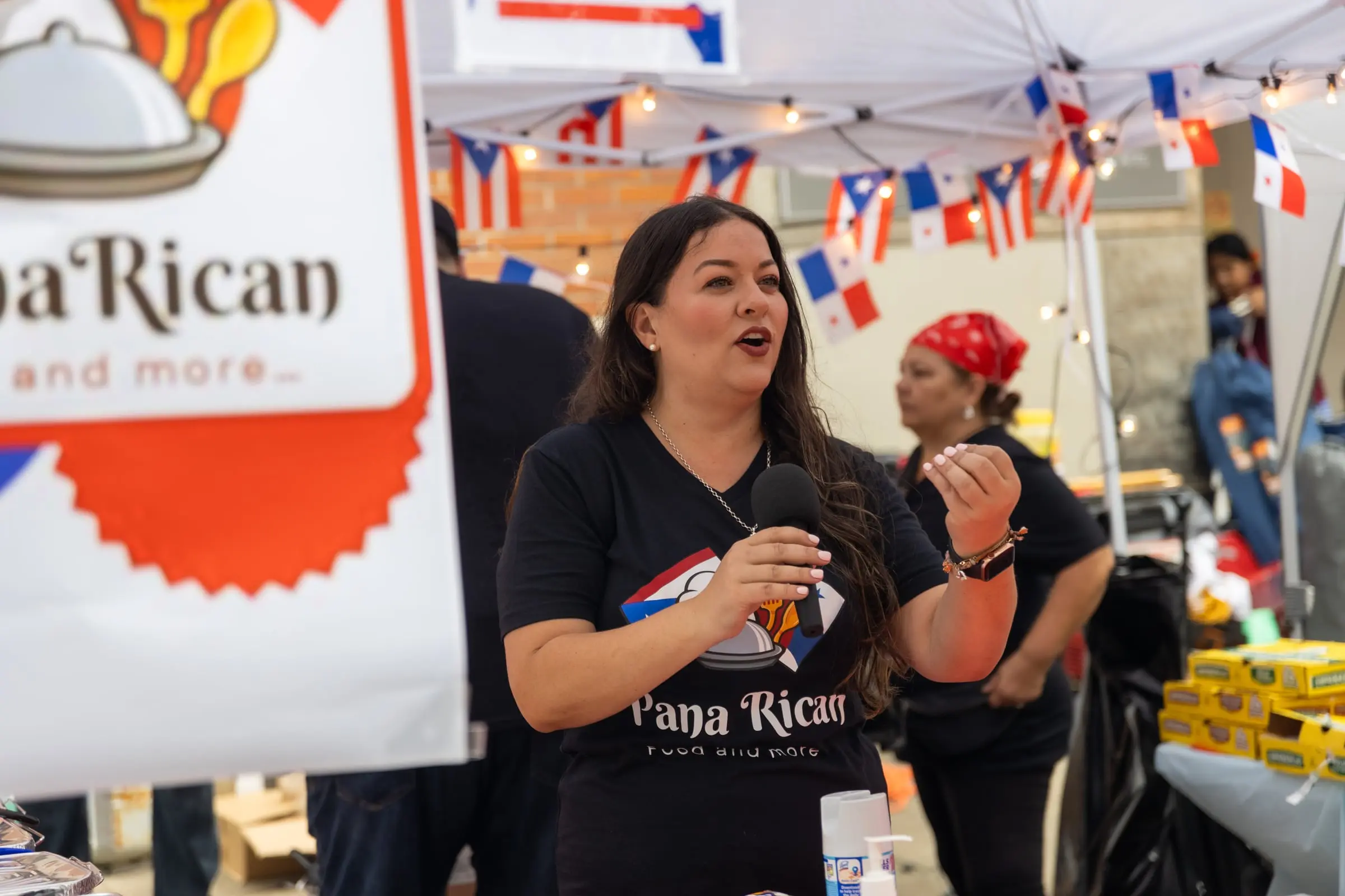 A Pana Rican vendor speaks into a microphone in front of her food stand, photographed by AVO Dynamics at Mobile Latin Fest 2023