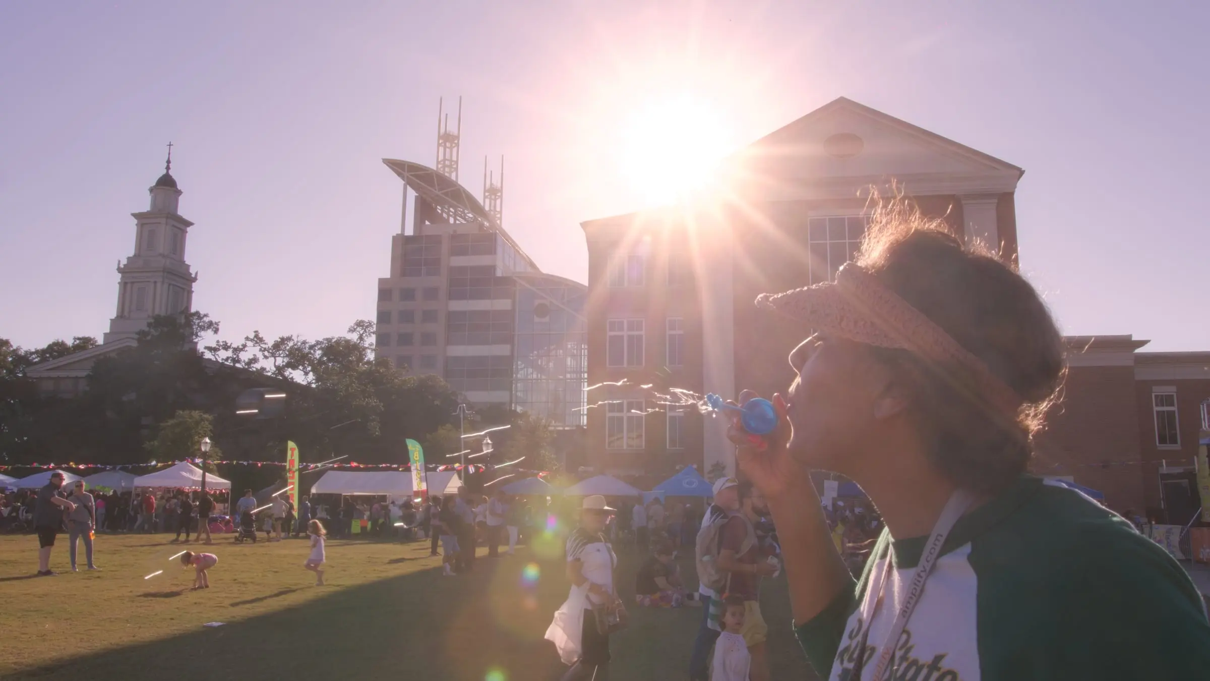 A festival-goer plays a whistle in late sun with the vendor tents and a clock tower behind, photographed by AVO Dynamics at Mobile Latin Fest 2023