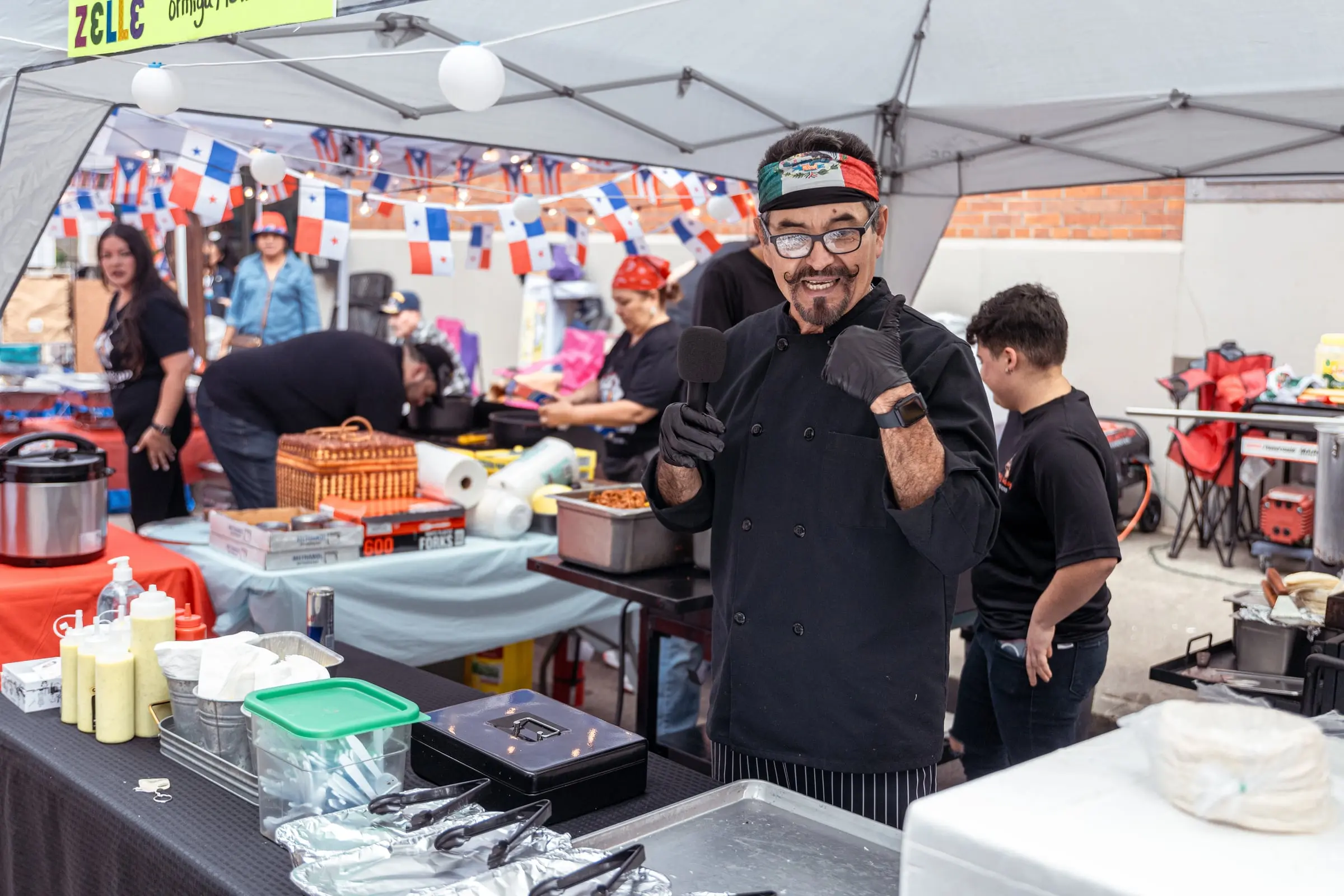 A food vendor's chef gives a thumbs-up at his tent while his crew cooks behind him, photographed by AVO Dynamics at Mobile Latin Fest 2023